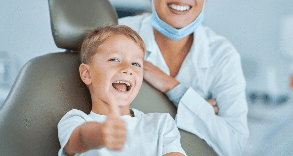 A happy young boy giving a thumbs up in a dental chair with a smiling dentist in the background.